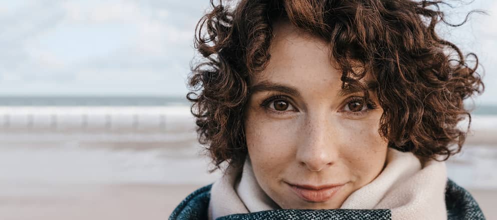 Vrouw staat met warme sjaal op het strand en kijkt de camera in